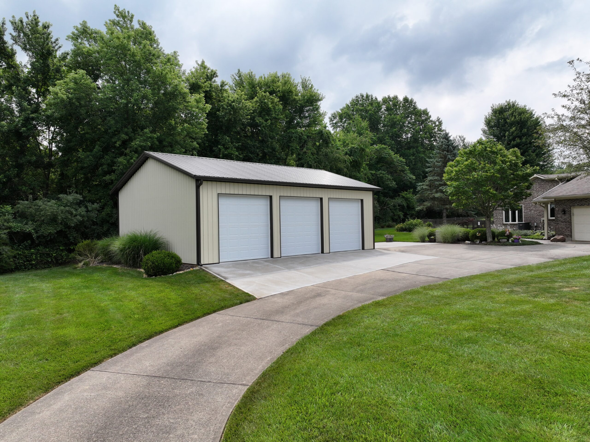 Three car post frame garage with tan siding and dark trim beside a brick home in Southern Ohio